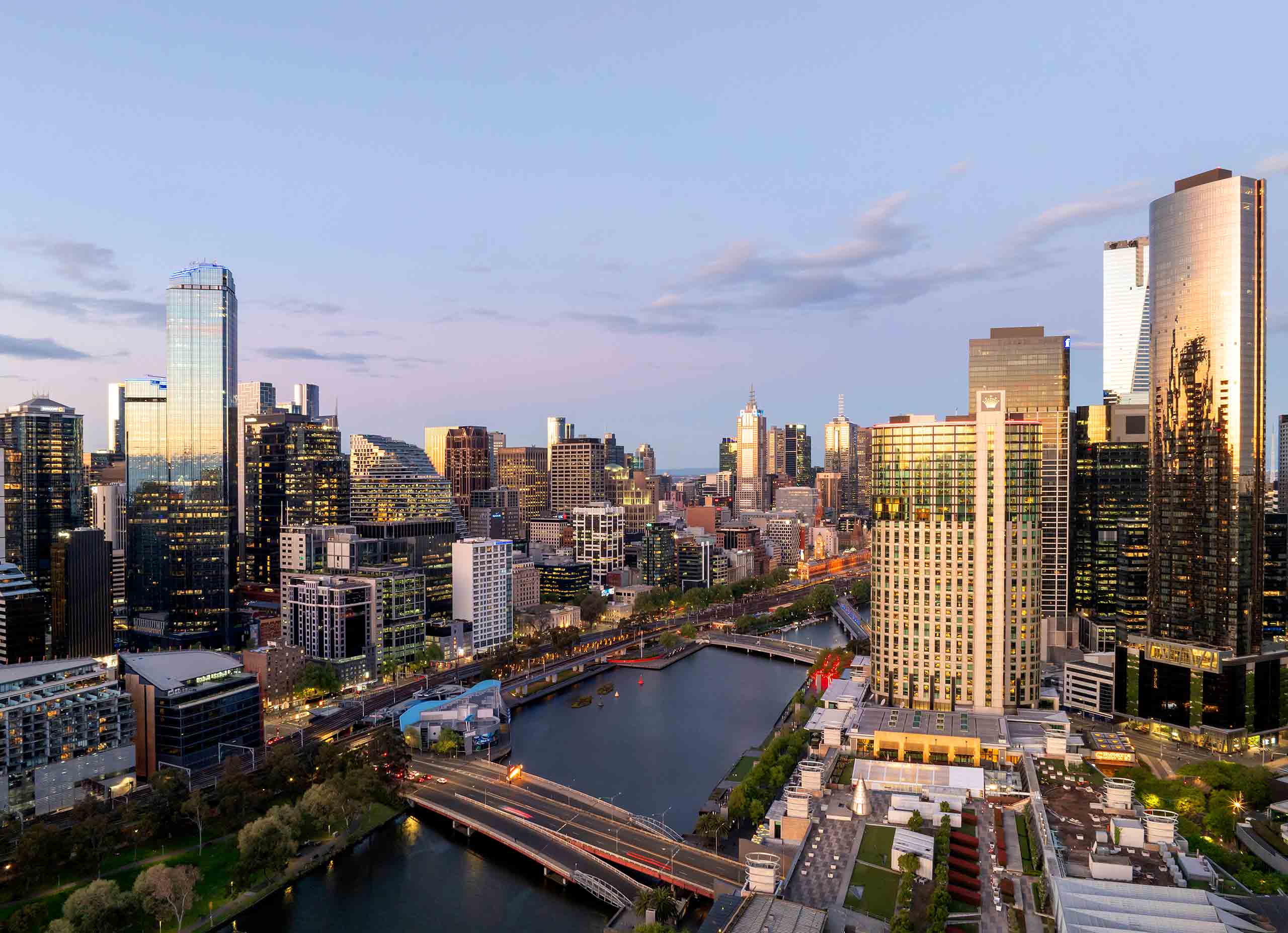Drone view of Melbourne skyline at dusk showing illuminated skyscrapers and Yarra River reflecting city lights.